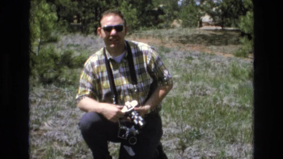Adult man in a yellow and green plaid shirt and sunglasses crouches in a grassy Lake Tahoe park area while holding a camera with a strap.