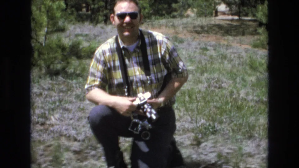 Smiling adult man with sunglasses holds a vintage camera while crouching among grass and low vegetation in the Lake Tahoe park setting.