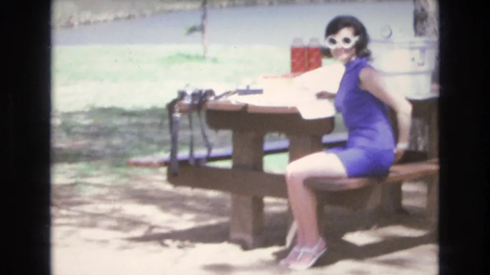 Adult woman in a blue sleeveless dress and white sunglasses sits at an outdoor picnic table with food and drink items in the supplied Lake Tahoe 1973 vacation footage.
