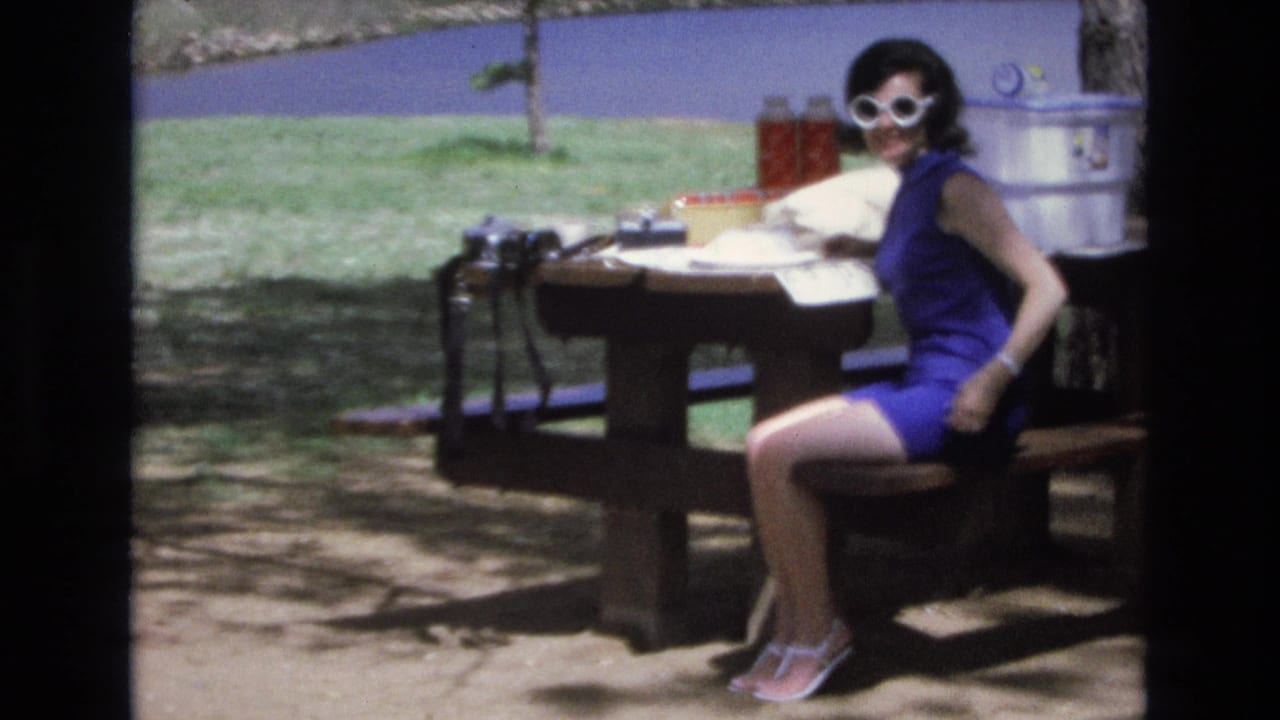 In Lake Tahoe, Nevada, a woman in a blue sleeveless dress and white sunglasses sits at a wooden picnic table with picnic gear and lake water behind her, consistent with the supplied 1973 home-movie metadata.