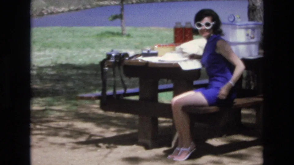 In Lake Tahoe, Nevada, a woman in a blue sleeveless dress and white sunglasses sits at a wooden picnic table with picnic gear and lake water behind her, consistent with the supplied 1973 home-movie metadata.