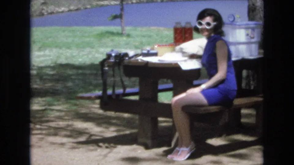 A woman in a blue dress and white sunglasses faces the camera from a lakeside picnic table, with a camera, thermos, cooler, grass, and lake water visible in the soft 8mm frame.