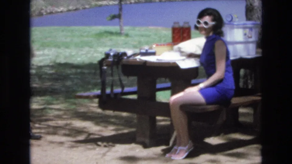 The woman sits in profile at the Lake Tahoe picnic table with her white sunglasses and blue dress visible, while the 8mm frame includes a dark vertical edge and lakeside grass and water in the background.