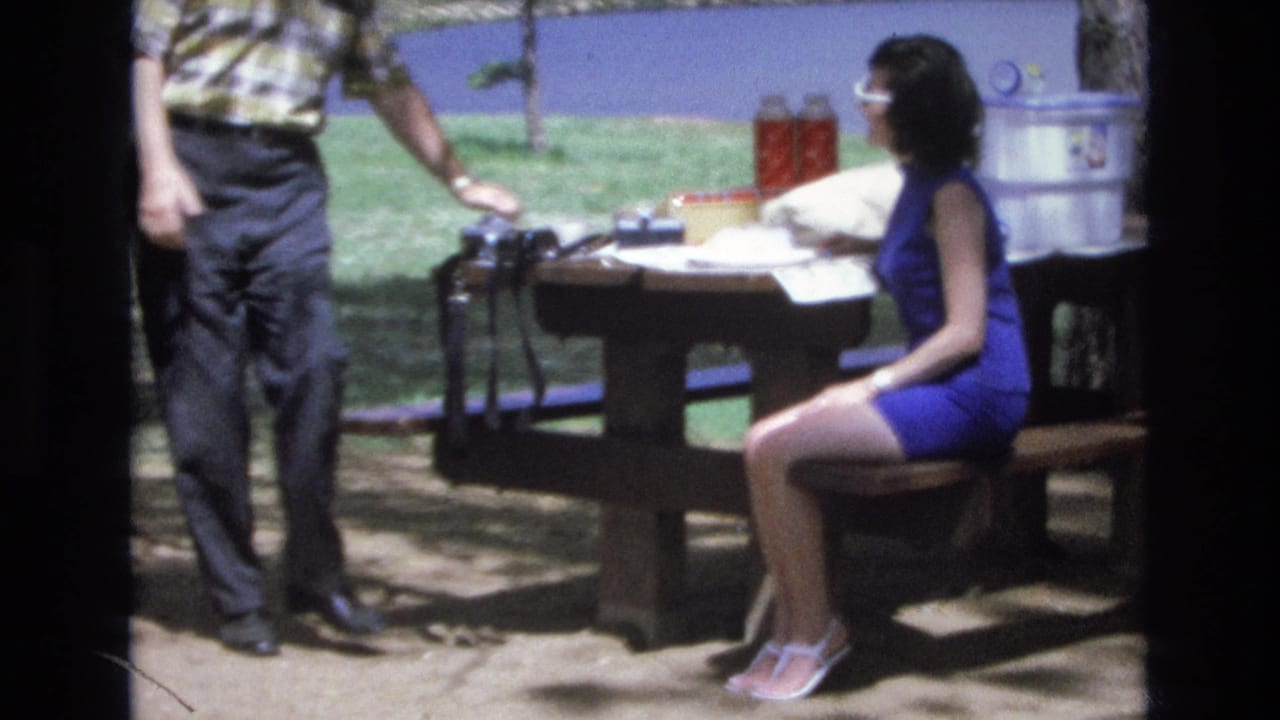 Man standing beside a wooden picnic table while a woman in a blue dress sits on the bench near a lake.