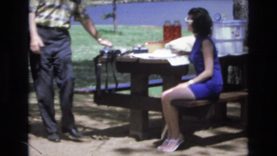 In the supplied 1973 Lake Tahoe, Nevada home-movie footage, a man stands near a wooden picnic table as a woman in white sunglasses and a blue dress sits beside picnic items with lake water behind them.