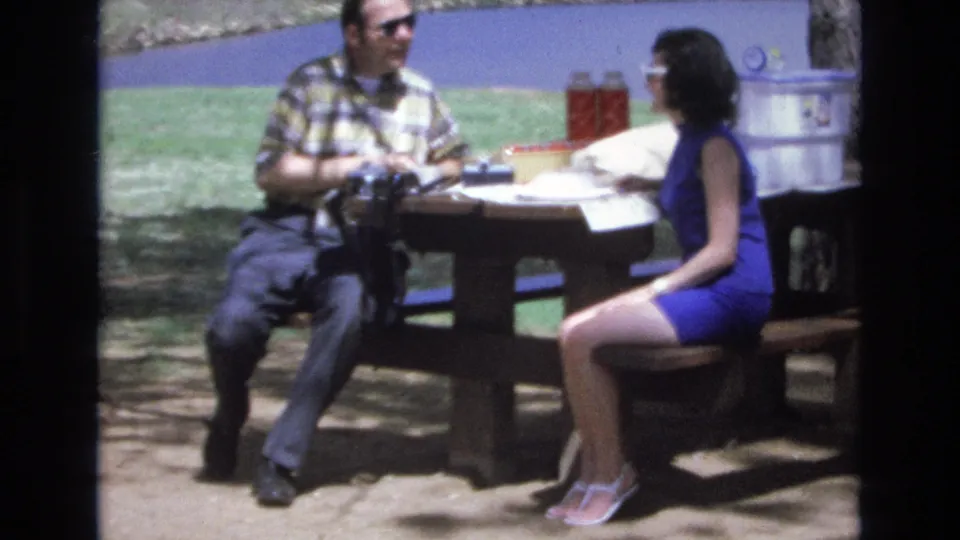 A seated couple face each other at the picnic table in the 1970s home-movie frame, with the man holding or handling an object near his lap and the woman turned toward him in conversation.
