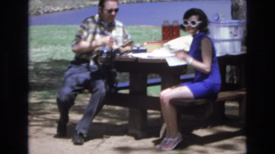 In the closing frame, the woman in a blue dress and white sunglasses turns toward the camera while seated at the Lake Tahoe picnic table, with the man, picnic supplies, trees, and lake water still visible.