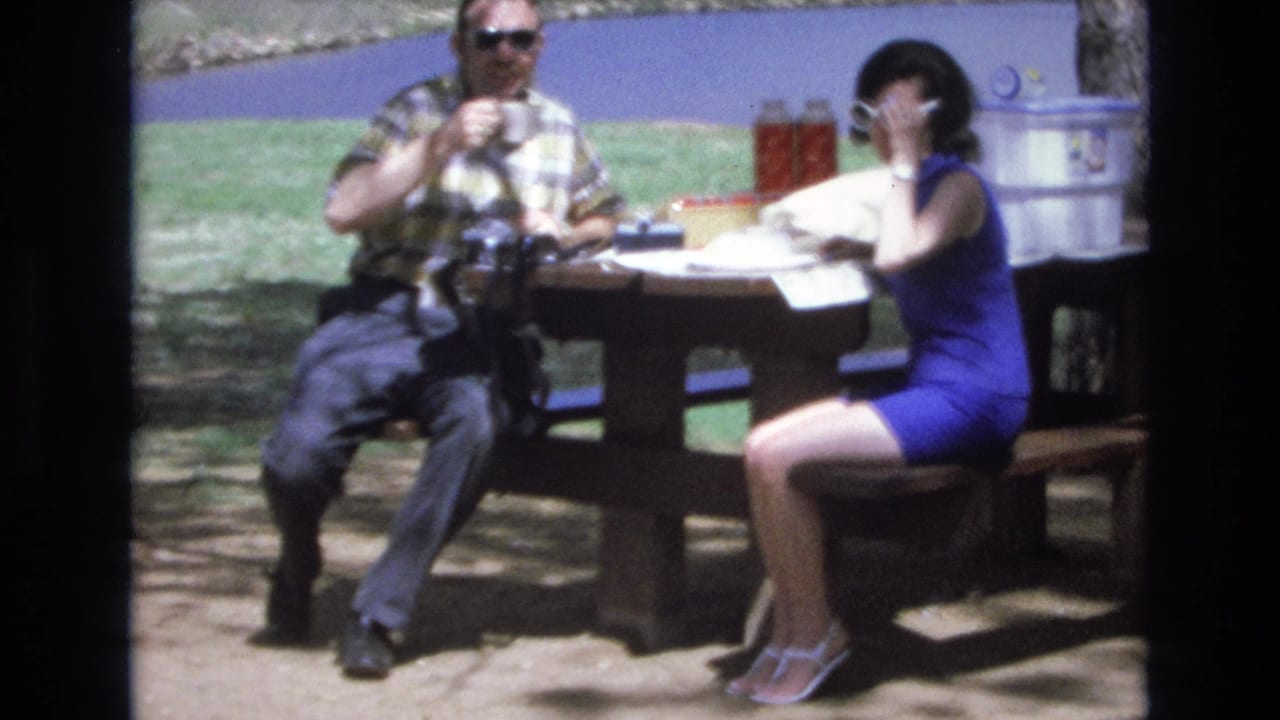 At Lake Tahoe, Nevada in 1973, a man in a plaid shirt and sunglasses and a woman in a blue dress sit at a wooden picnic table with drinks, papers, thermoses, and a cooler.