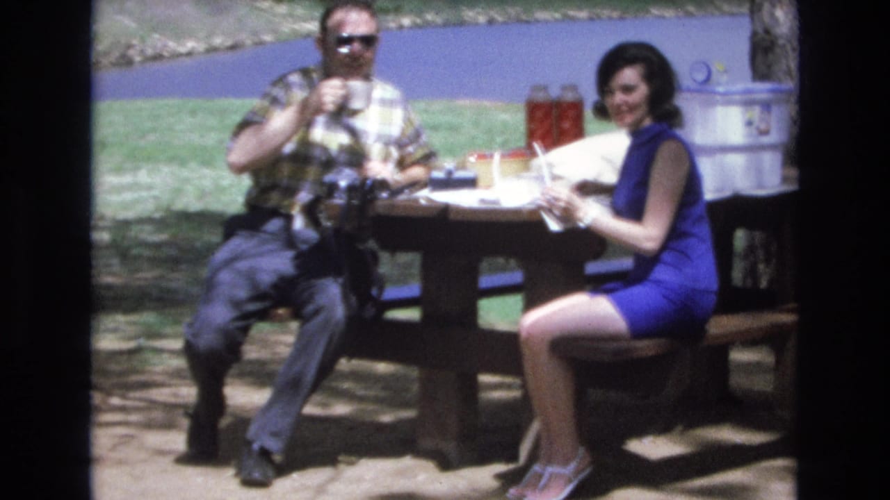 Woman smiles toward the camera while seated beside a man at a picnic table.