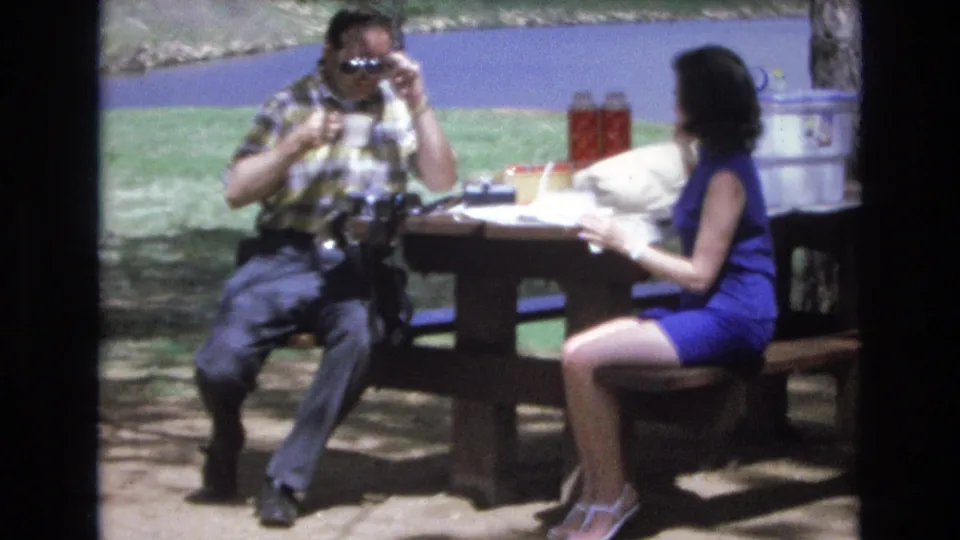 The man adjusts his sunglasses while seated at the picnic table, with the woman turned partly away and papers, cups, thermoses, grass, and water visible around them.