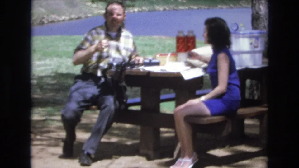 The two adults face one another at the Lake Tahoe picnic table, with the man holding a cup and the woman seated beside papers, red thermoses, a cooler, grass, and water.