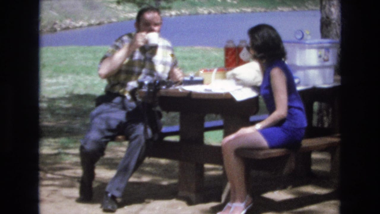 Man drinks from a mug while a woman sits across the picnic table.