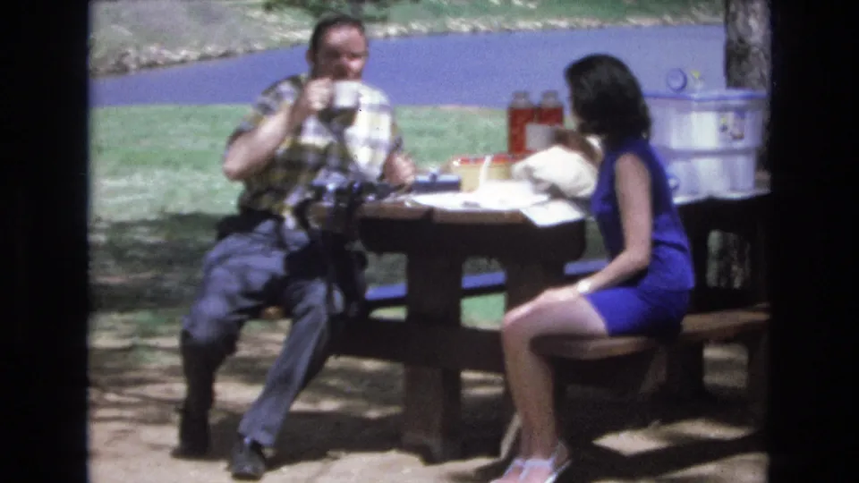 A home-movie frame shows the couple in conversation at the picnic table, with the man holding a mug and the woman leaning slightly forward near food, drinks, and a large cooler.