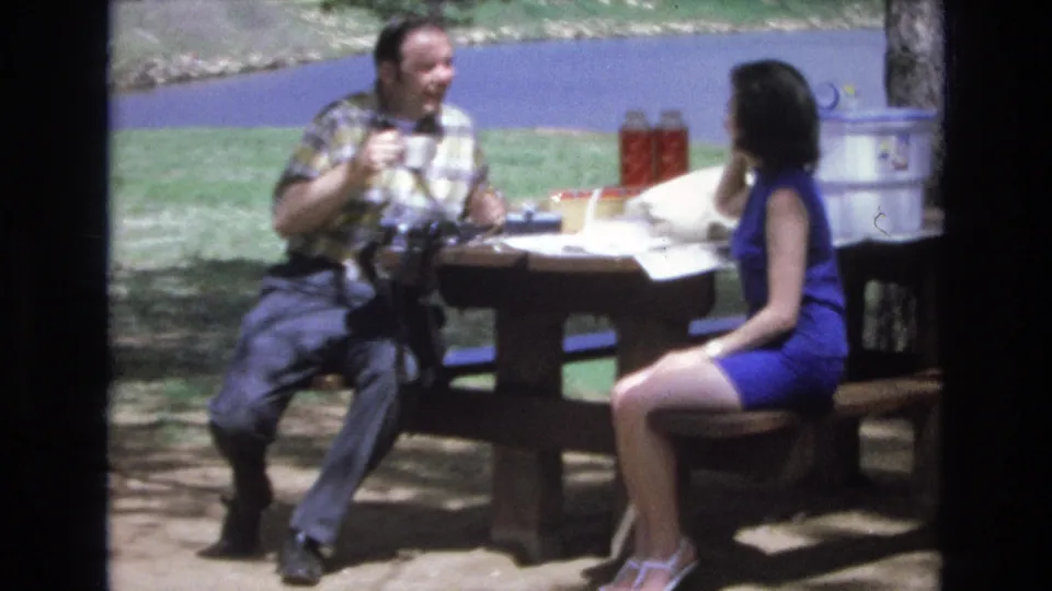 The final frame keeps the couple at the lakeside picnic table, with the man holding a mug and the woman turned toward him beside picnic supplies and a large cooler.