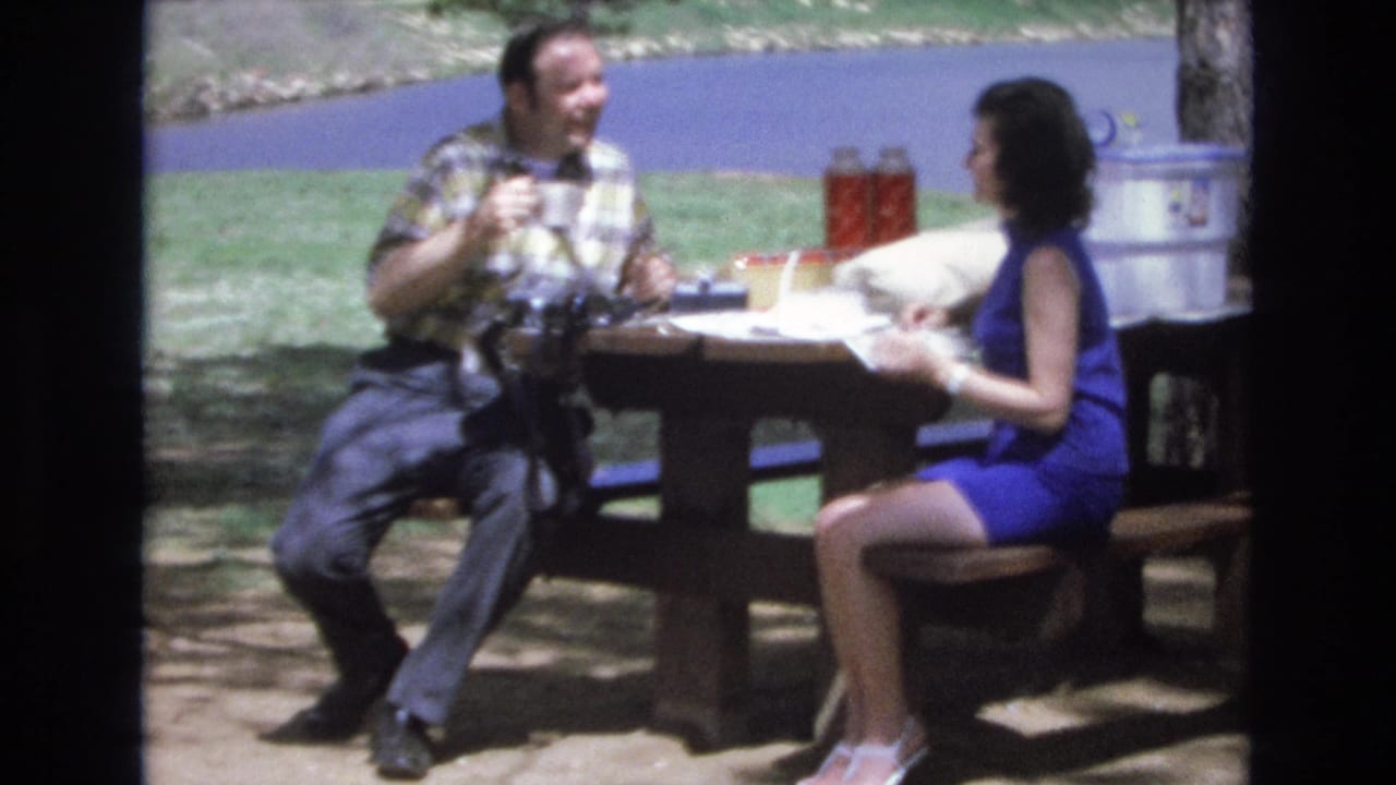 In 1973 Lake Tahoe, Nevada home-movie footage, a man in a plaid shirt and a woman in a blue dress sit at a picnic table with drinks and picnic items nearby.