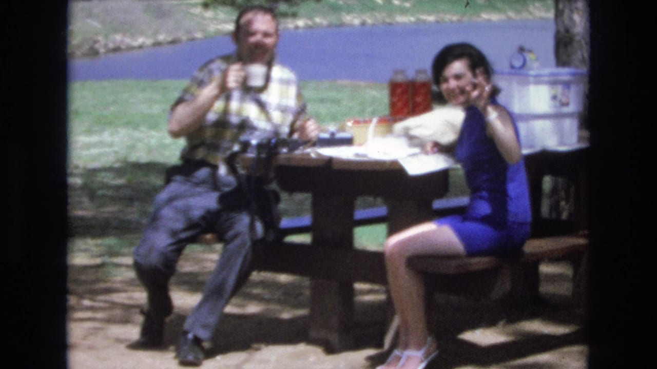 Woman at a picnic table waves toward the camera while a man holds a cup.