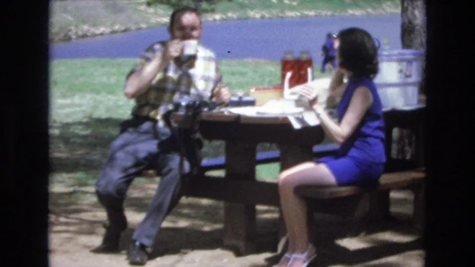 The man drinks from a cup as the woman turns toward the table holding a paper or wrapper, continuing the casual 1973 lakeside picnic scene.