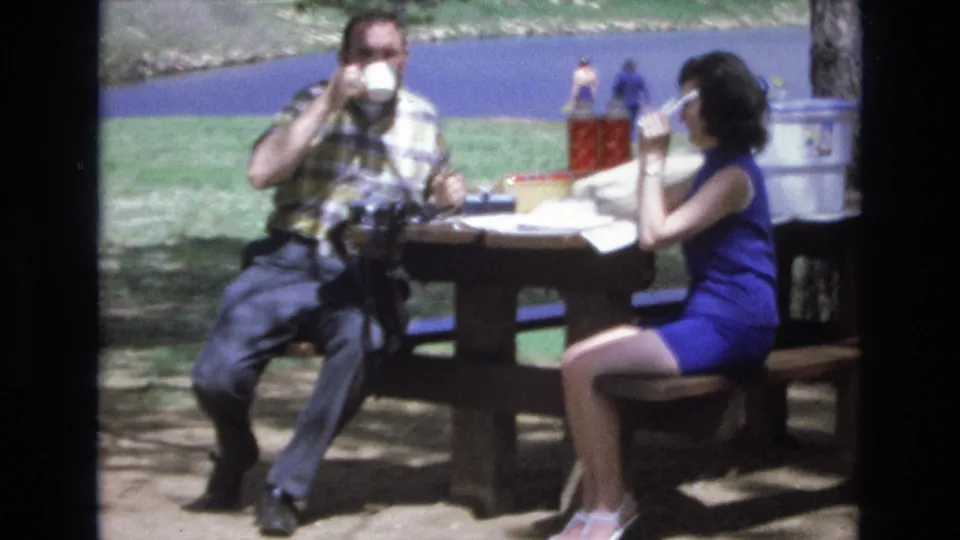 Both adults lift cups while seated at the wooden picnic table, with red drink containers, a cooler, grass, trees, and Lake Tahoe water visible in the background.