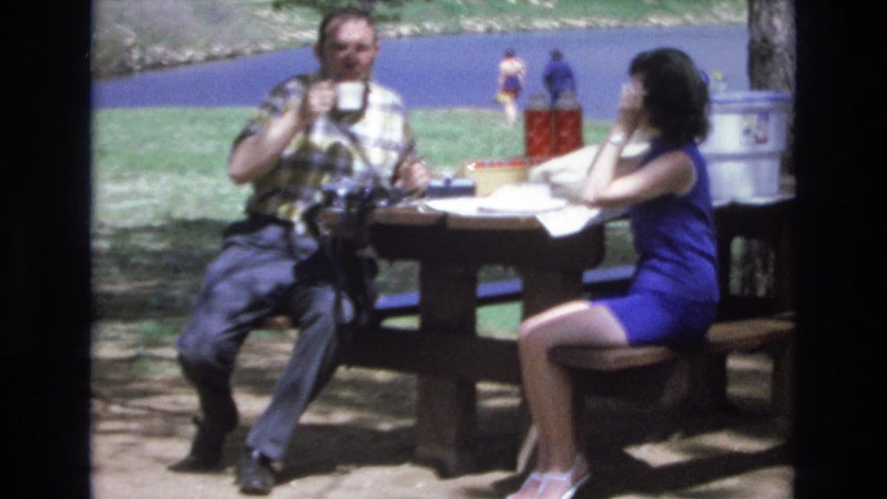 In supplied 1973 Lake Tahoe, Nevada home-movie footage, a man in a plaid shirt and a woman in a blue dress sit at a wooden picnic table near the water as the man holds a white cup.