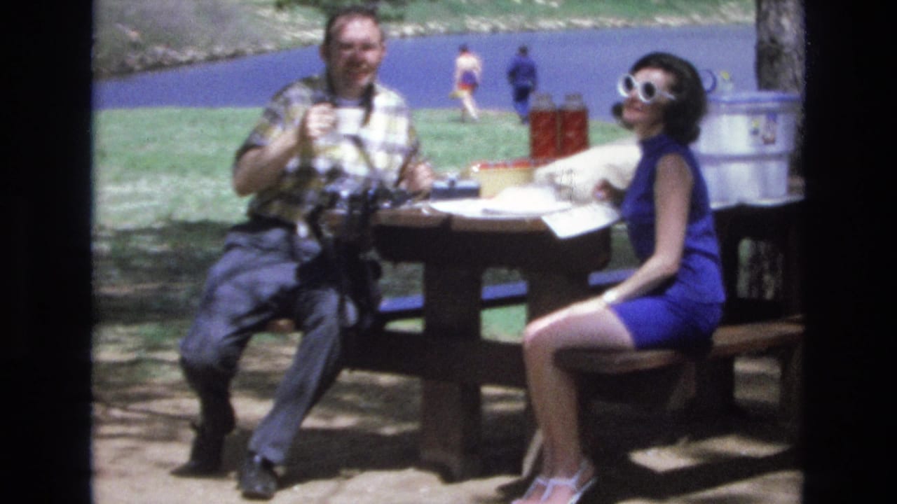 Woman in white sunglasses looks toward the camera as a man sits beside her at a picnic table.