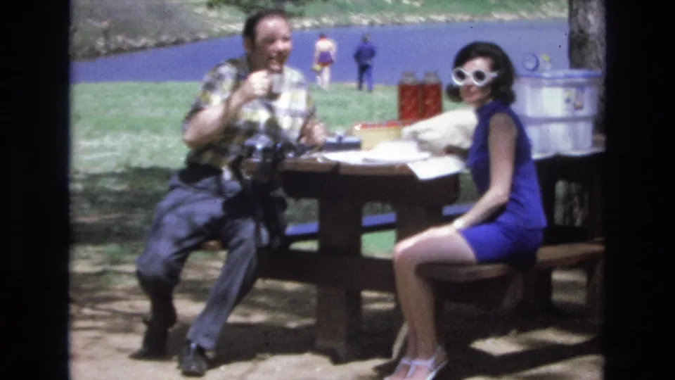 The couple appears to acknowledge the camera during a 1973 Lake Tahoe picnic, with the woman smiling in white sunglasses and the man seated beside cups and food items on the table.