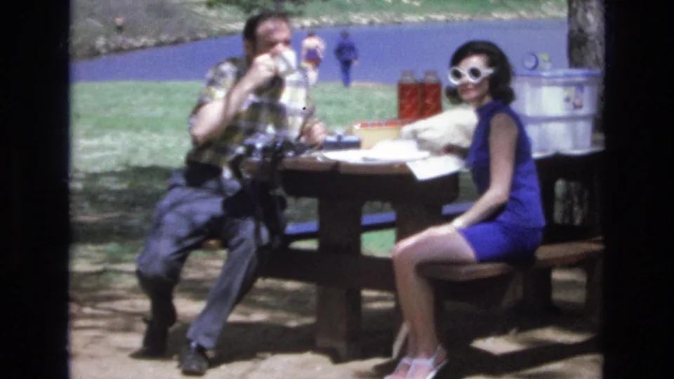 At the end of the clip, the man drinks from a cup while the woman in a blue dress and white sunglasses sits facing the camera at the Lake Tahoe picnic table.