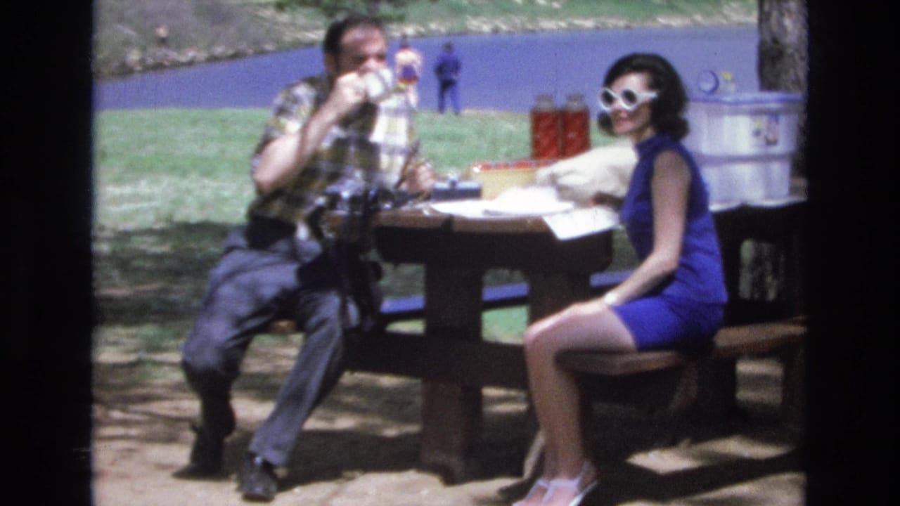 A man and woman sit at a wooden picnic table near lake water, with the man drinking from a cup.