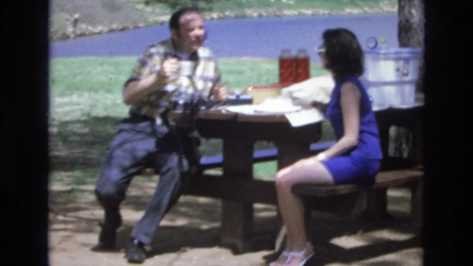 The couple remains seated at the lakeside wooden table in the supplied 1973 Lake Tahoe footage, with the man holding a cup and the woman listening across from him.
