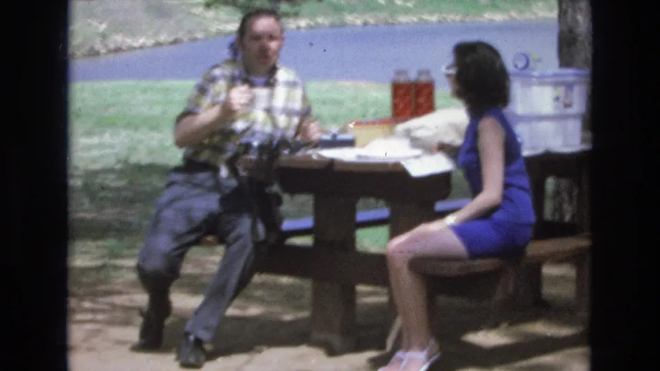 At the end of the clip, the man at the Lake Tahoe picnic table appears to look toward the camera while the woman sits beside him, with the cooler, grass, and water still visible.