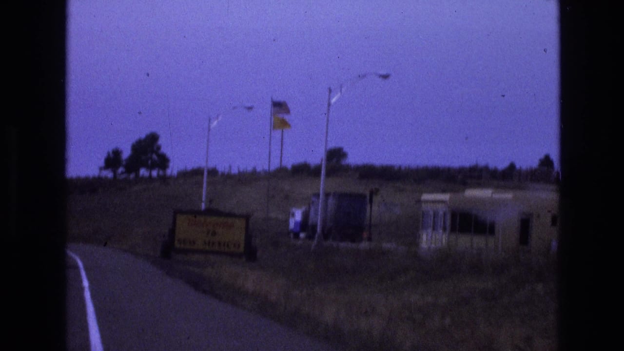 1973 8mm road-trip frame approaching a Welcome to New Mexico roadside sign, with flags, dry grass, a low hillside, and a small roadside structure visible through soft purple-tinted film color.