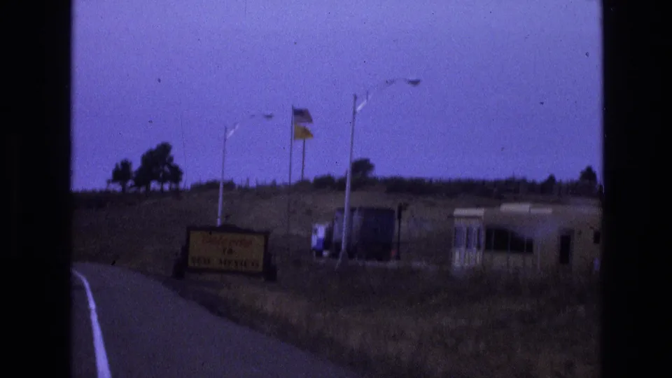 1973 8mm road-trip frame approaching a Welcome to New Mexico roadside sign, with flags, dry grass, a low hillside, and a small roadside structure visible through soft purple-tinted film color.