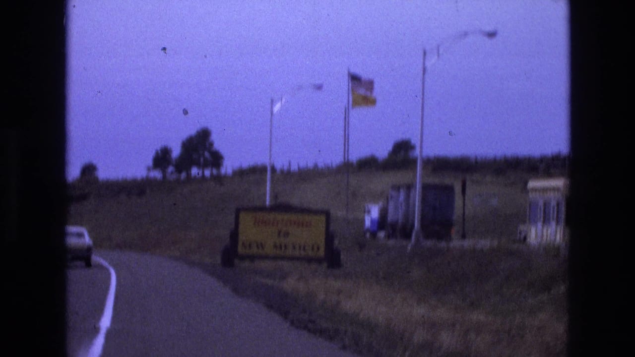 1973 home-movie frame from a moving car showing the Welcome to New Mexico sign on the roadside, flags above it, a white vehicle ahead, and small structures to the right.