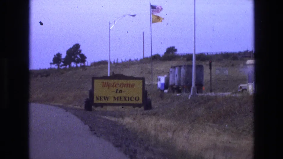 1973 New Mexico road-trip still with the yellow Welcome to New Mexico sign more legible, U.S. and state flags above, a road edge at left, and utility or roadside structures on the right.