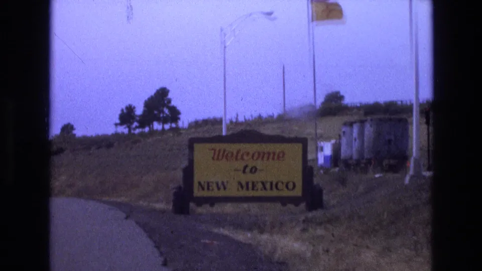 Final 1973 8mm frame with the Welcome to New Mexico sign near the foreground, flags overhead, a low grassy hillside, light poles, and small roadside structures in the background.