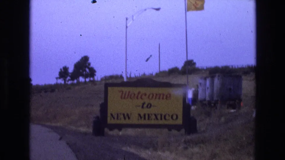 1973 8mm frame showing a Welcome to New Mexico roadside sign near a road, with dry hillside terrain, streetlights, and film gate edges visible.