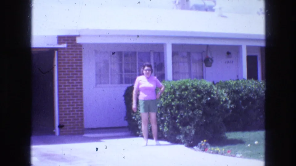 1973 8mm frame showing a woman in a pink top and green shorts standing in a driveway outside a single-story suburban house with hedges and a carport or garage area.