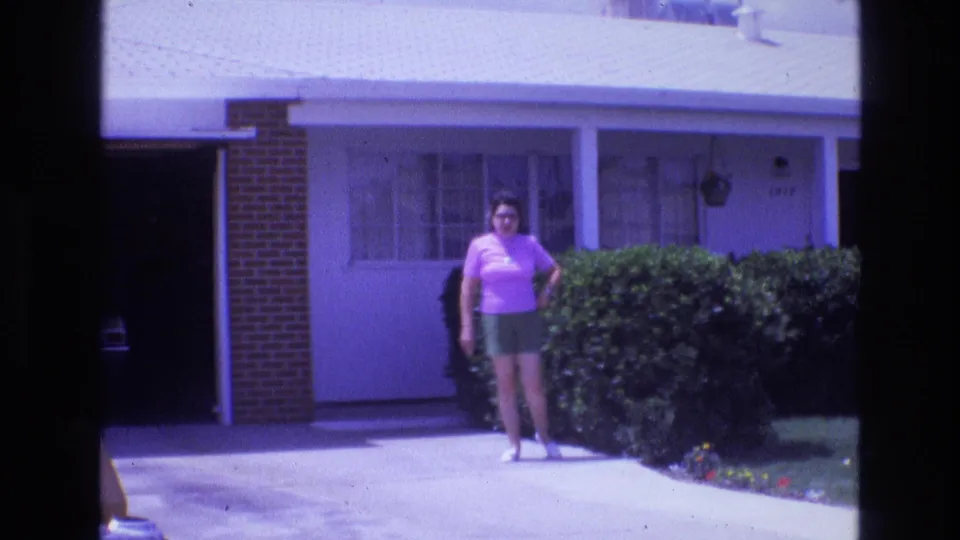 1973 home-movie frame of a woman posing near the driveway and hedge of a single-story suburban house, with the image softened by exposure and purple-blue film color shift.