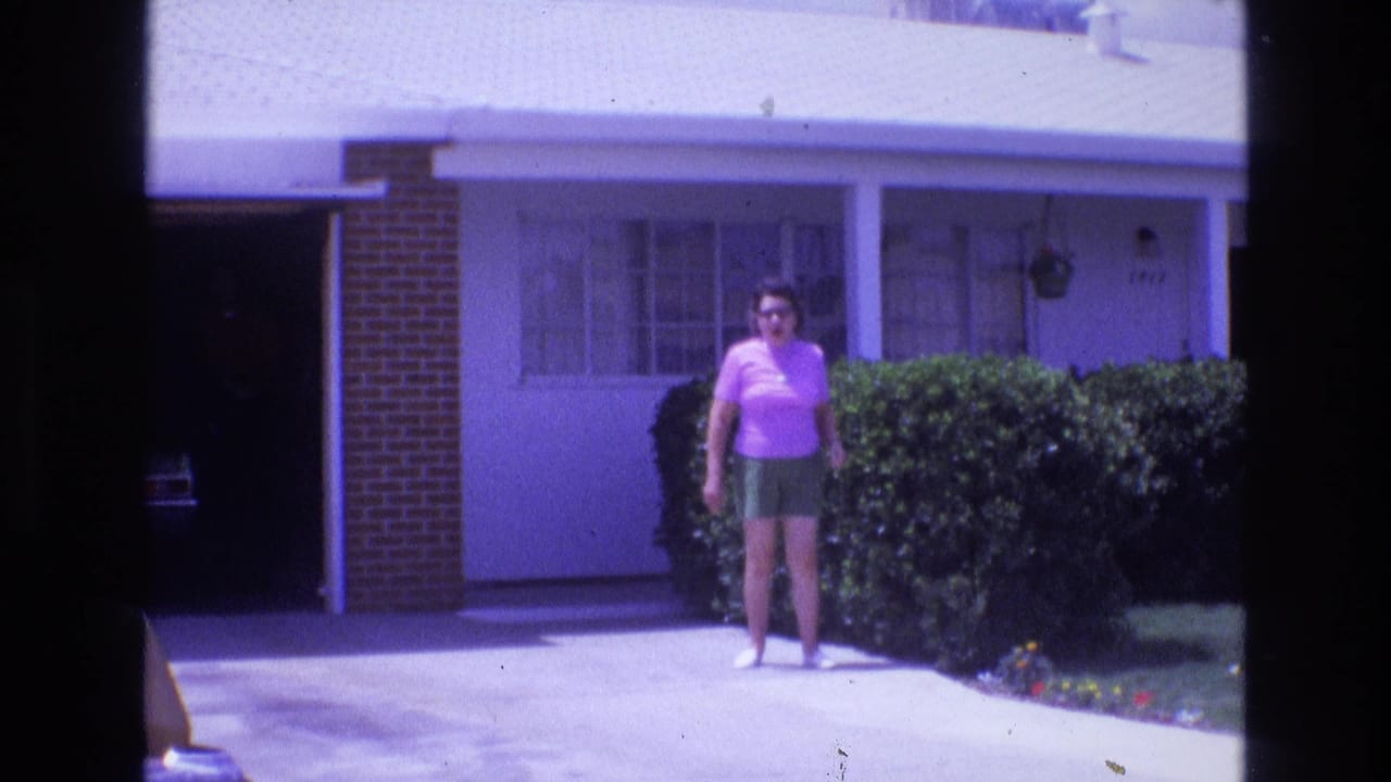 1973 New Mexico 8mm frame showing an adult woman in a pink shirt and green shorts near a driveway, with a garage, front windows, and hedges behind her.