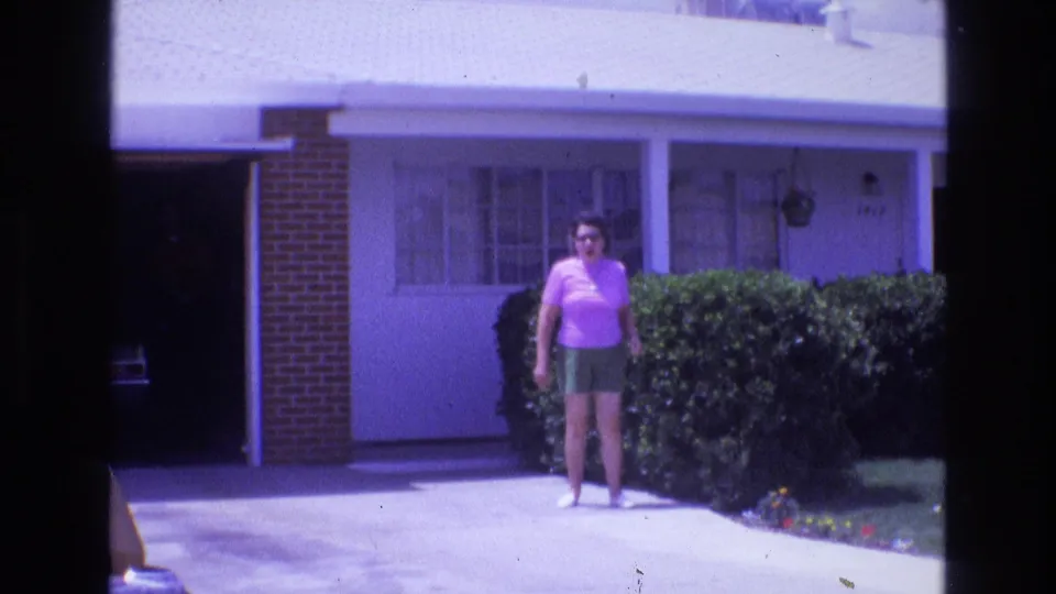1973 New Mexico 8mm frame showing an adult woman in a pink shirt and green shorts near a driveway, with a garage, front windows, and hedges behind her.