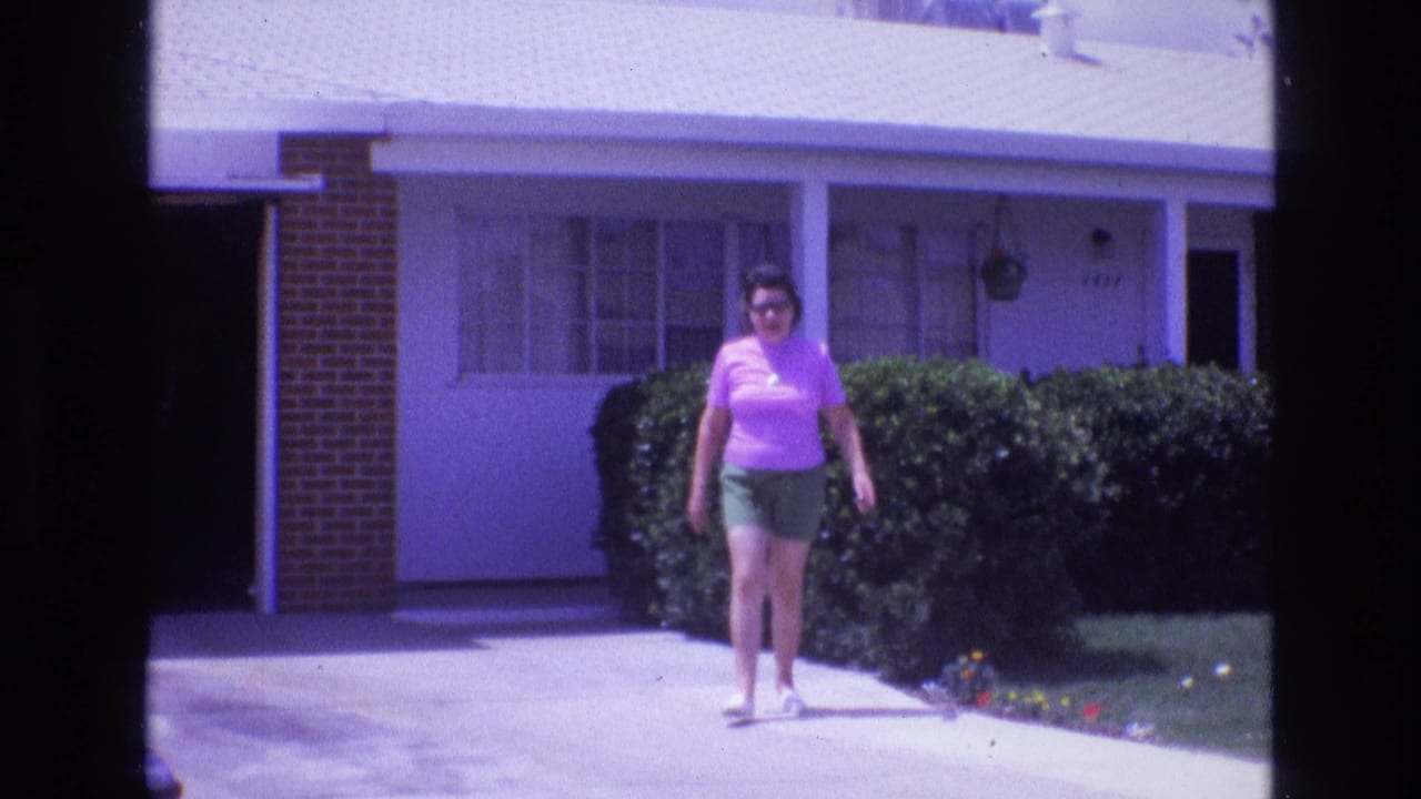 1973 New Mexico home-movie frame of an adult woman walking toward the camera along a concrete driveway outside a single-story residential house.