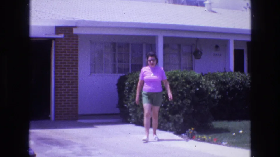 1973 New Mexico home-movie frame of an adult woman walking toward the camera along a concrete driveway outside a single-story residential house.