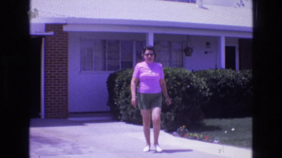 1973 New Mexico 8mm frame showing an adult woman in casual summer clothing walking closer to the camera, with a residential driveway, garage, and trimmed bushes in view.