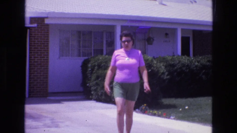 Close 1973 New Mexico home-movie frame of an adult woman in sunglasses walking down a driveway, with hedges, lawn, flowers, and the house exterior behind her.