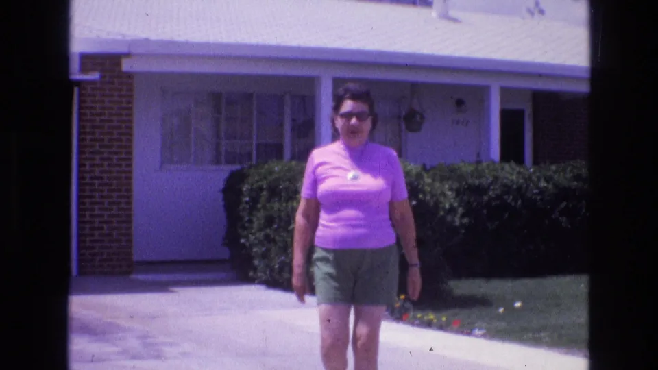 In New Mexico home-movie footage from 1973, an adult woman in a pink shirt and green shorts walks toward the camera beside a suburban driveway and hedges.