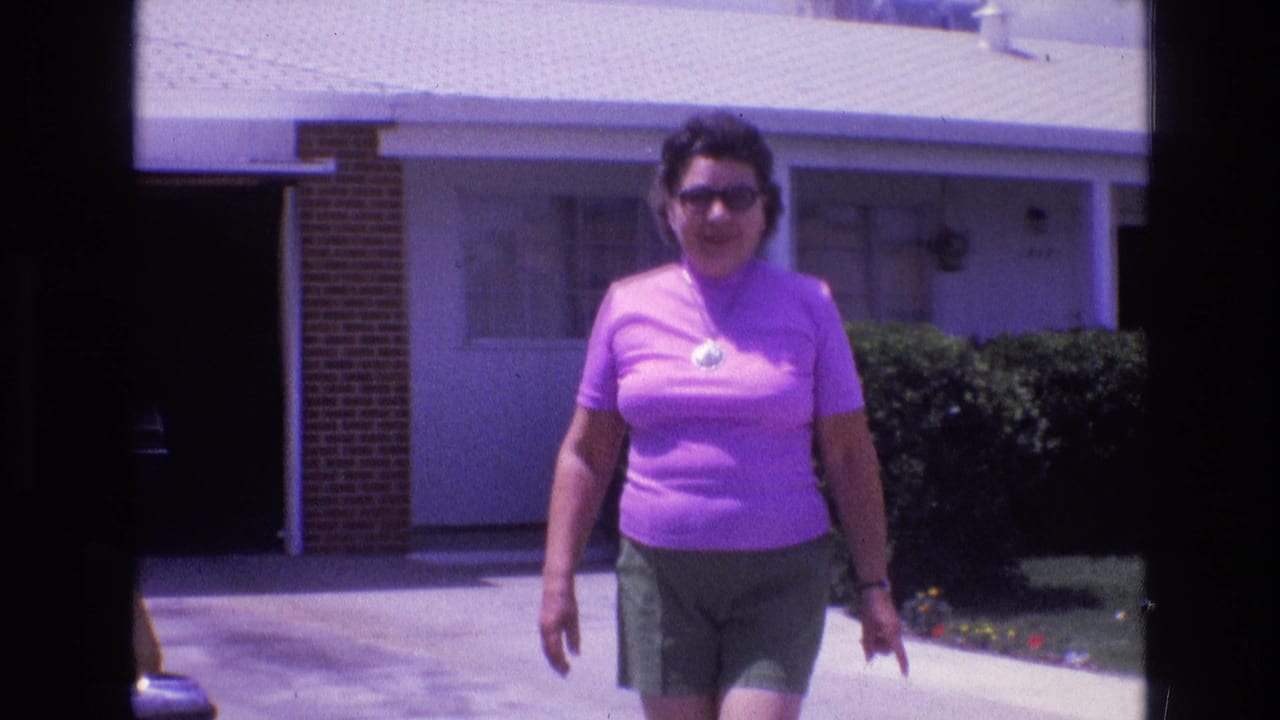 Woman in a pink shirt approaches the camera with a house and hedges behind her.