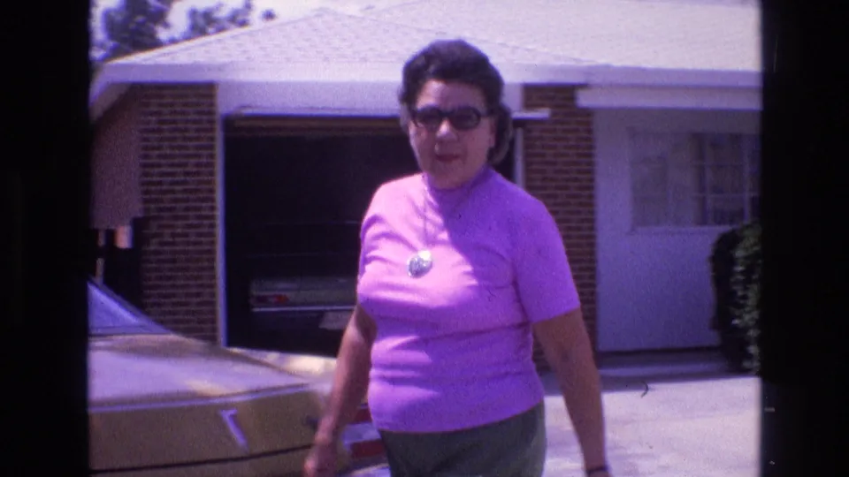 The adult woman passes closer to the lens, with a gold-colored car, open garage area, driveway, and single-story house visible behind her.