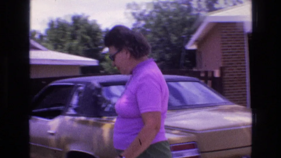 A side view shows the woman walking past a gold-colored car in the driveway, with neighboring roofs and trees visible in the background.
