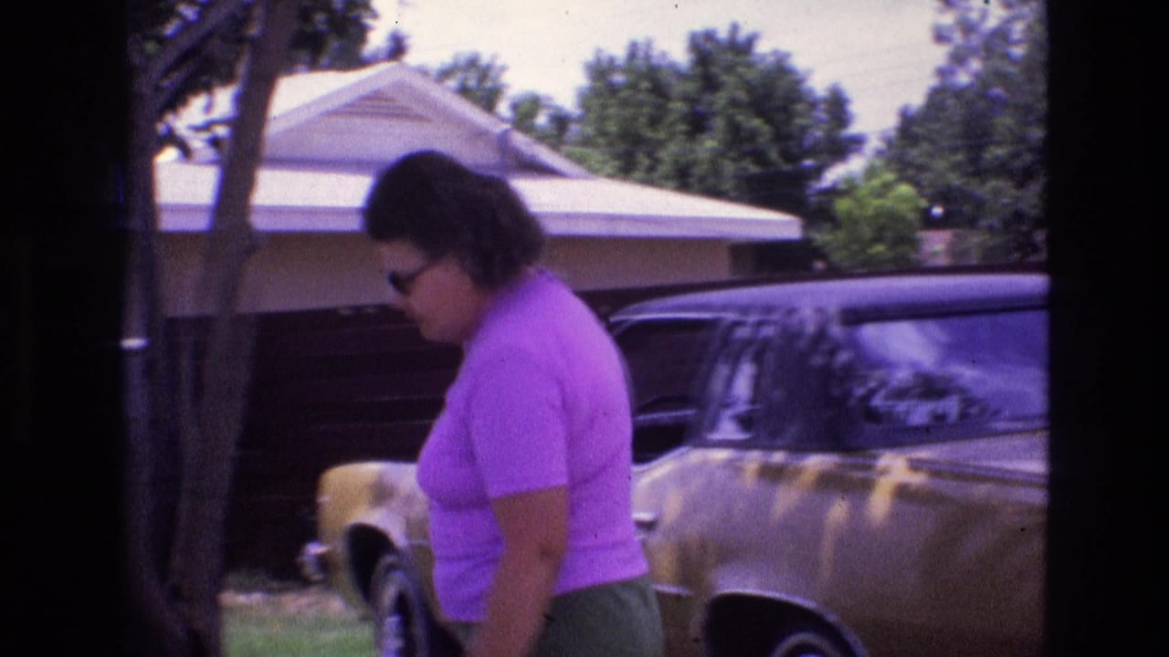 In a 1973 New Mexico home-movie frame, a woman in a purple shirt and green shorts bends near a gold-colored parked car outside a single-story suburban house.