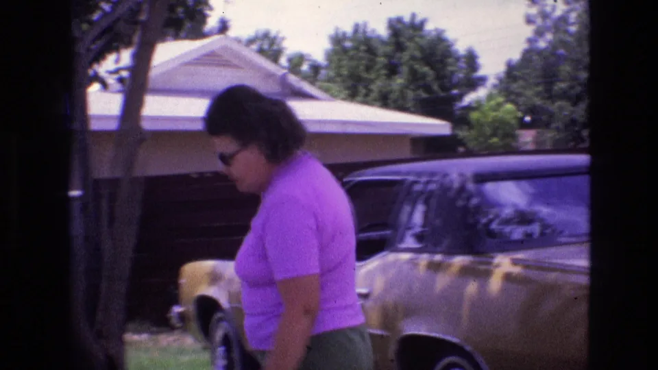In a 1973 New Mexico home-movie frame, a woman in a purple shirt and green shorts bends near a gold-colored parked car outside a single-story suburban house.