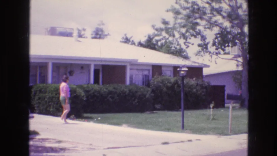 The woman appears farther from the camera, walking near the curb or driveway in front of hedges and a single-story house in 1973 New Mexico home-movie footage.